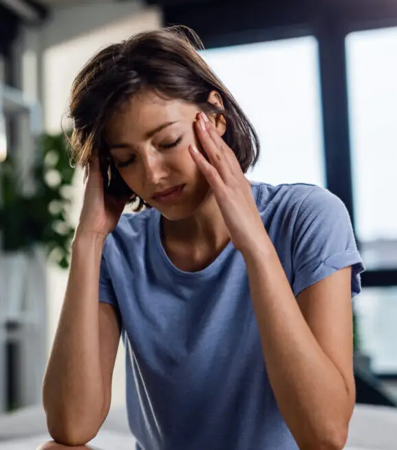 Sad woman suffering from headache and sitting on the bed with eyes closed while holding her head in pain.